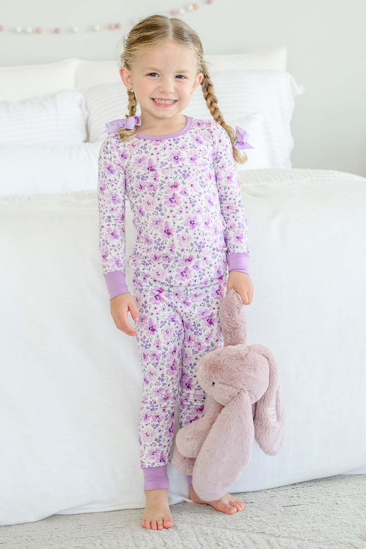 A grinning girl with braided pigtails standing by a bed wearing a Caden Lane cream knit pajama set in a Lilac Dreams floral print, paired with a plush bunny.