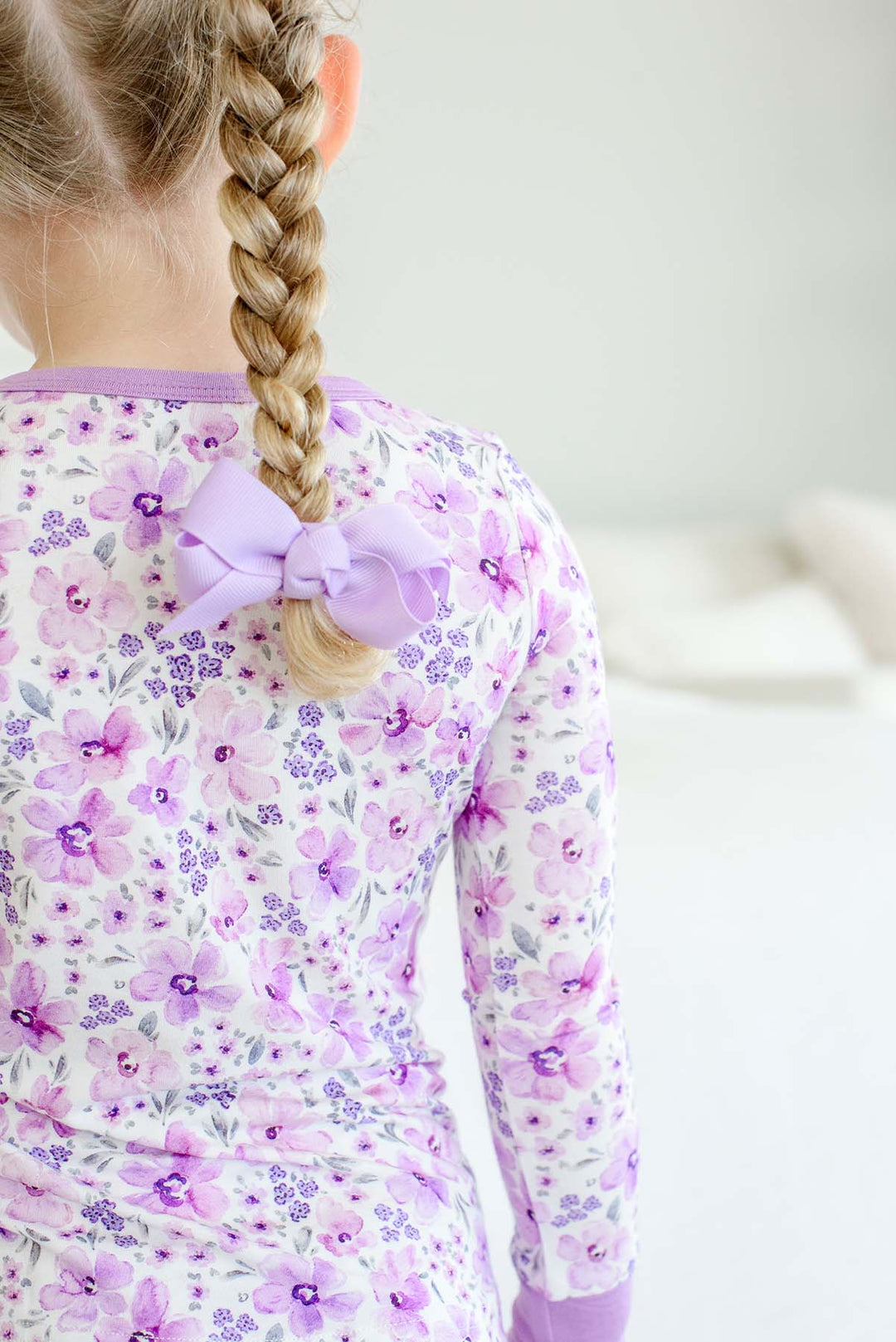 Back view of a girl with a lilac-bowed braid wearing a Caden Lane cream knit pajama top in a Lilac Dreams print featuring watercolor and tiny purple flowers.