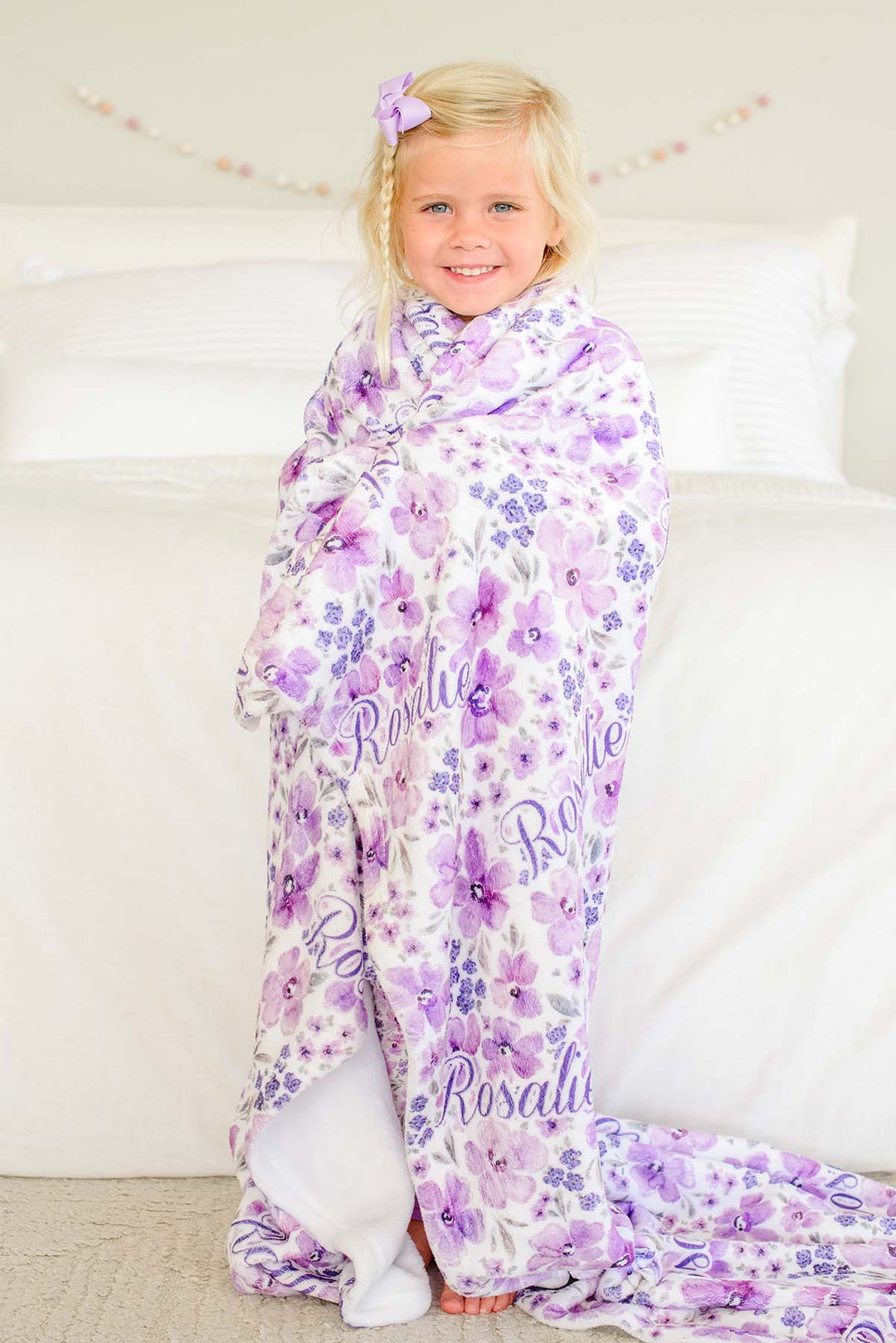 Full-length shot of a smiling toddler with a lilac bow, wrapped in a Caden Lane white plush blanket in a Lilac Dreams print featuring purple watercolor blooms, reading "Rosalie."