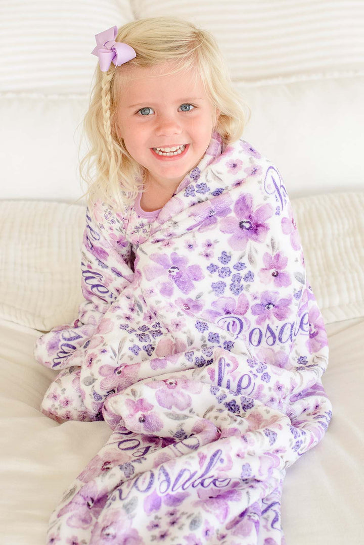 Grinning toddler seated on a bed, bundled up to her chin in a Caden Lane white plush blanket in a Lilac Dreams print featuring lavender florals, reading "Rosalie."