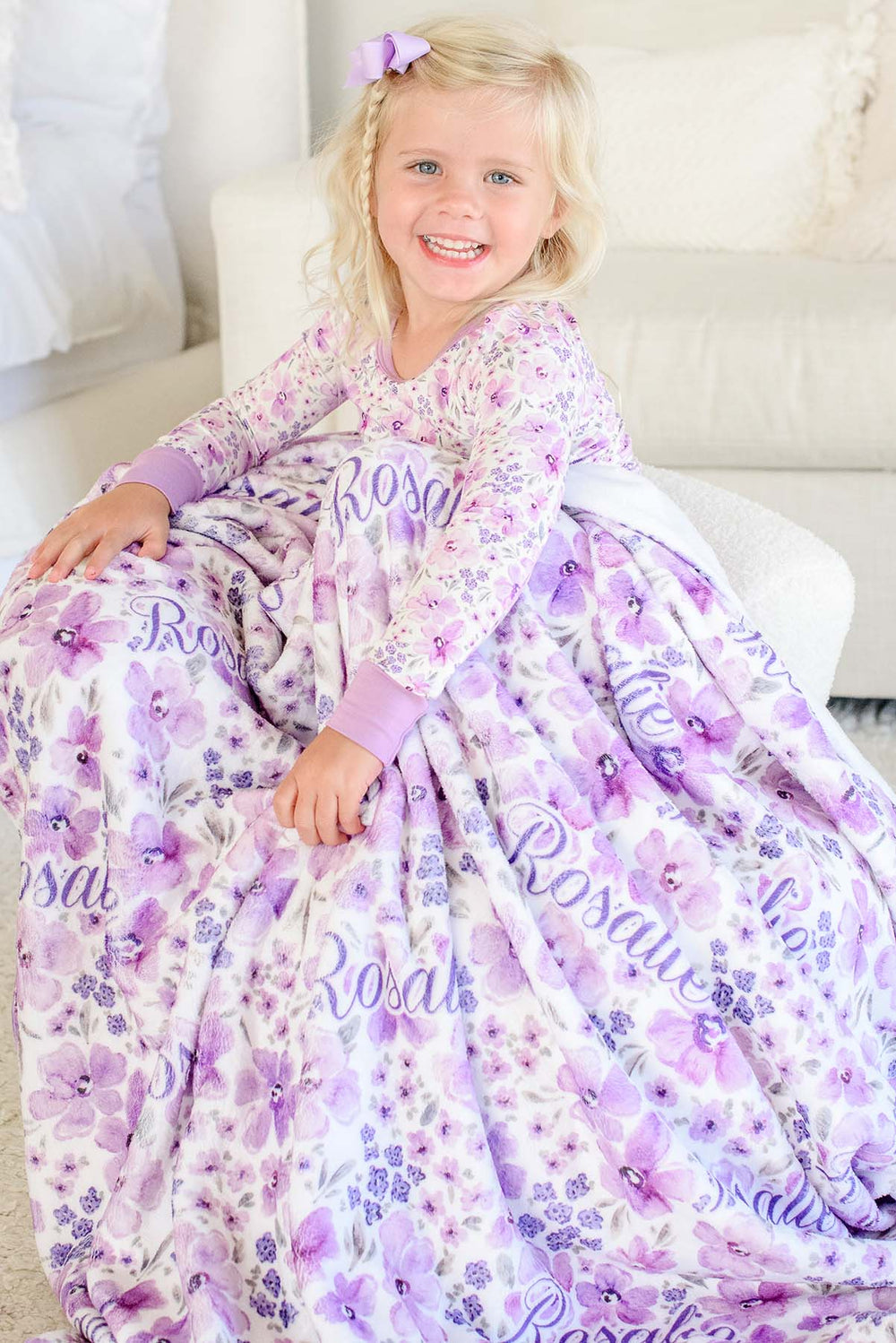 Laughing toddler on a white bouclé seat under a Caden Lane white plush blanket in a Lilac Dreams print featuring purple blooms, reading "Rosalie," paired with matching floral pajamas.