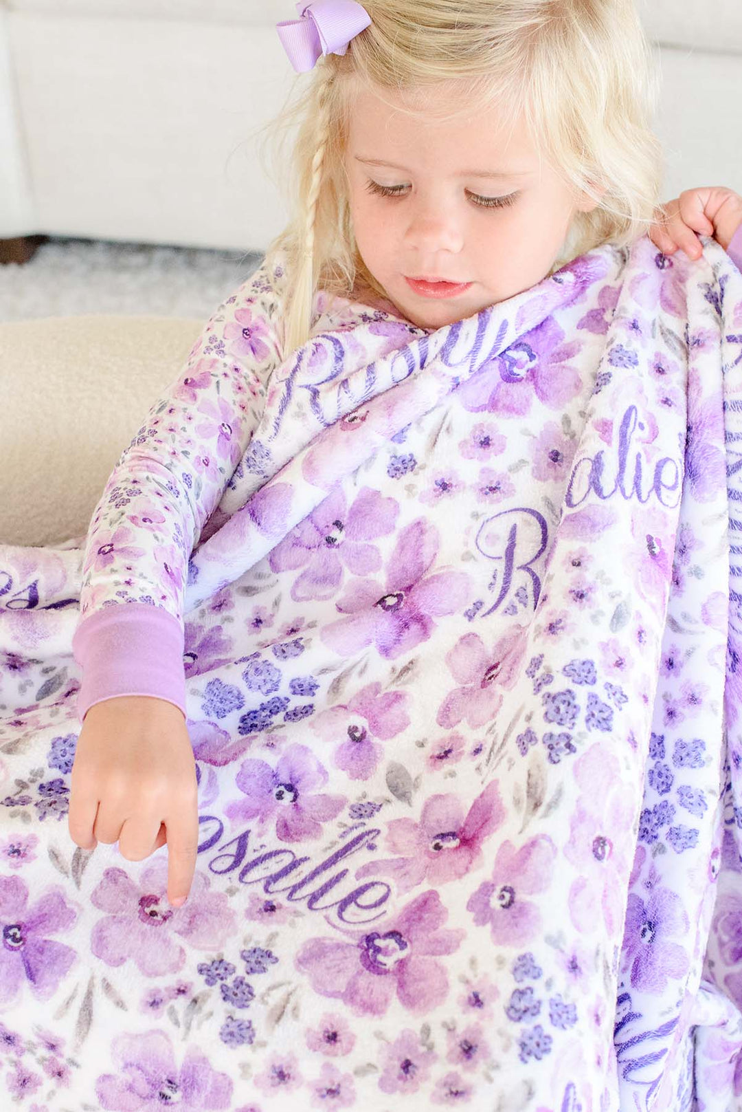 Close-up of a toddler pointing to a flower on a Caden Lane white plush blanket in a Lilac Dreams print featuring purple blooms, reading "Rosalie," paired with matching floral pajamas.