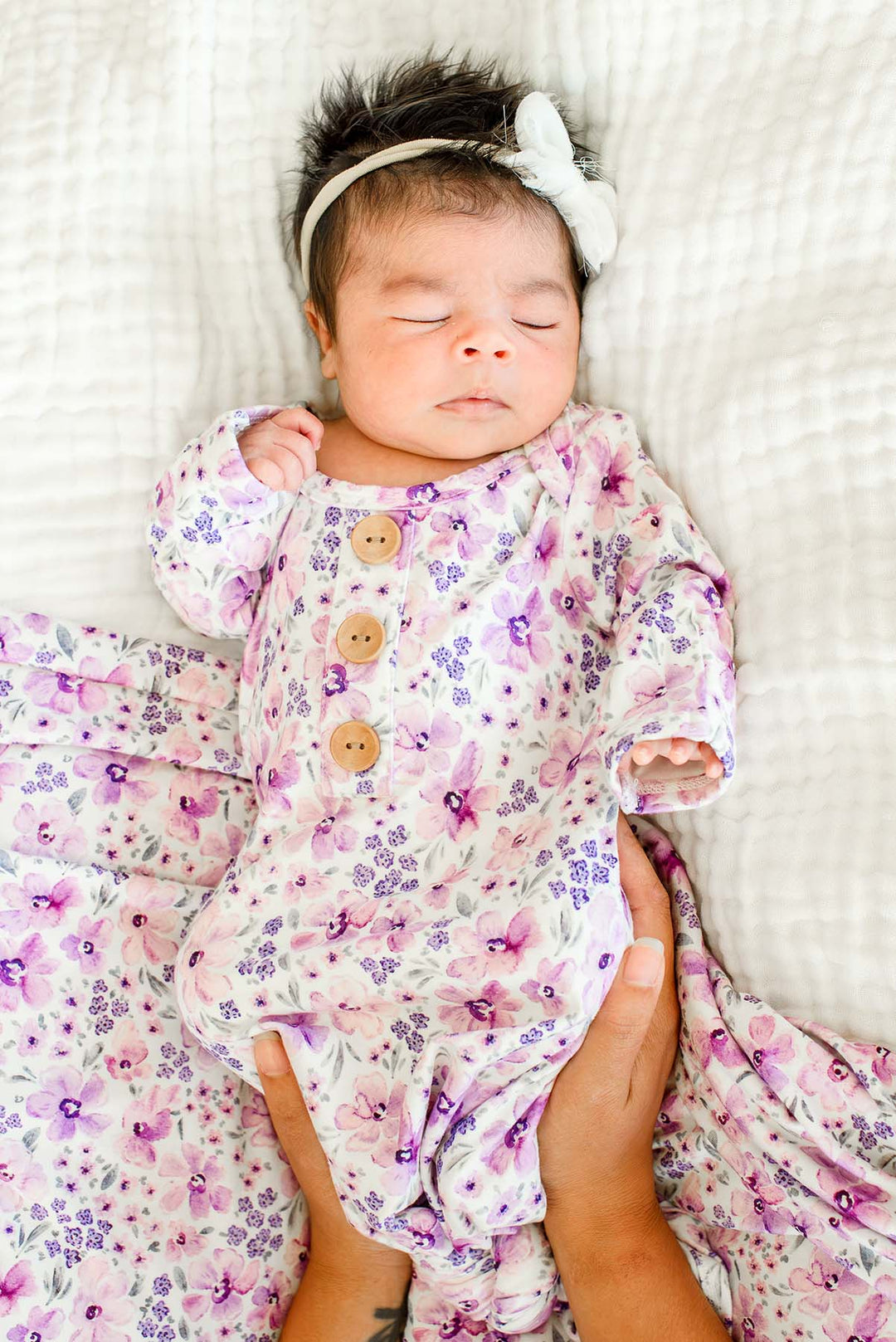 Sleeping newborn cradled by an adult's hands, wearing a Caden Lane white knit knotted gown in a Lilac Dreams print featuring purple flowers, paired with a matching swaddle.