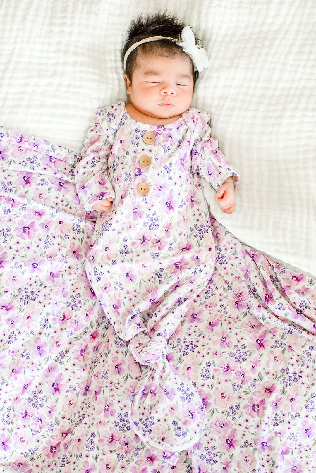 Full-length view of a sleeping newborn wearing a Caden Lane white knit knotted gown with wooden buttons in a Lilac Dreams print featuring pink blooms, paired with a matching swaddle.