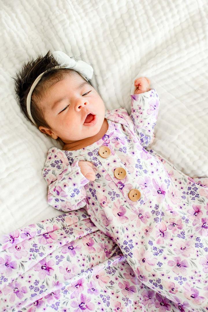 Angled shot of a newborn mid-coo wearing a Caden Lane white knit knotted gown with wooden buttons in a Lilac Dreams print featuring lavender florals, paired with a matching swaddle.