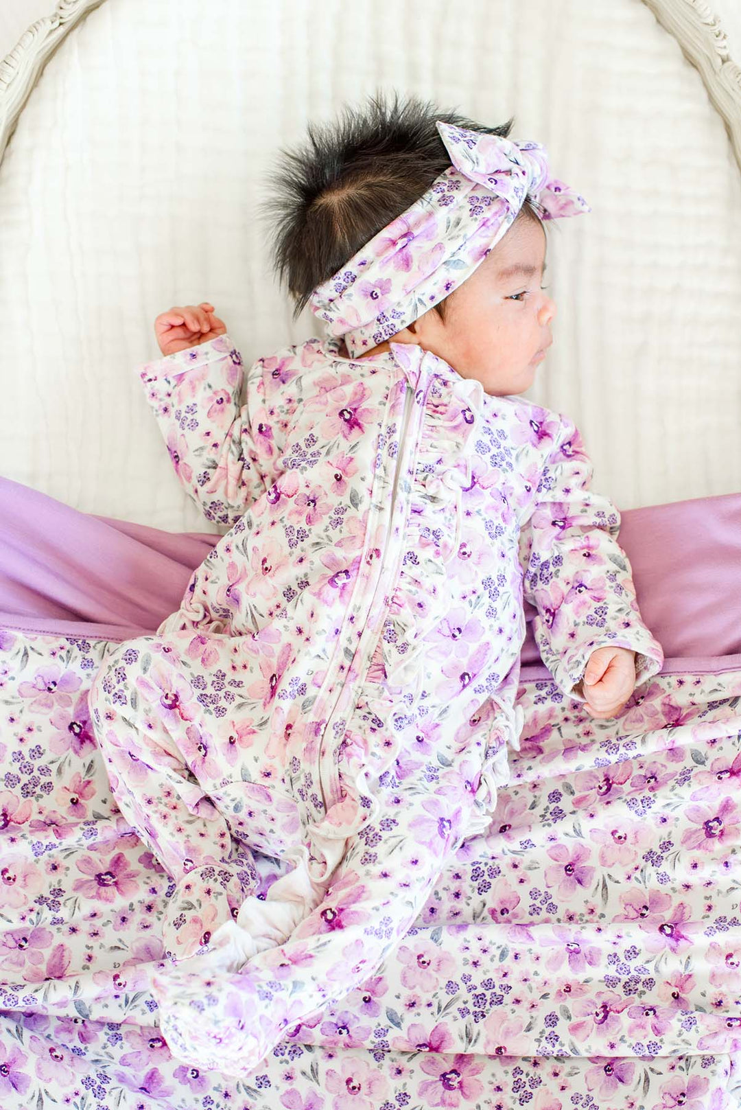 Side view of a newborn in a wicker bassinet wearing a Caden Lane white knit ruffle footie in a Lilac Dreams print featuring purple flowers, paired with a matching headwrap.