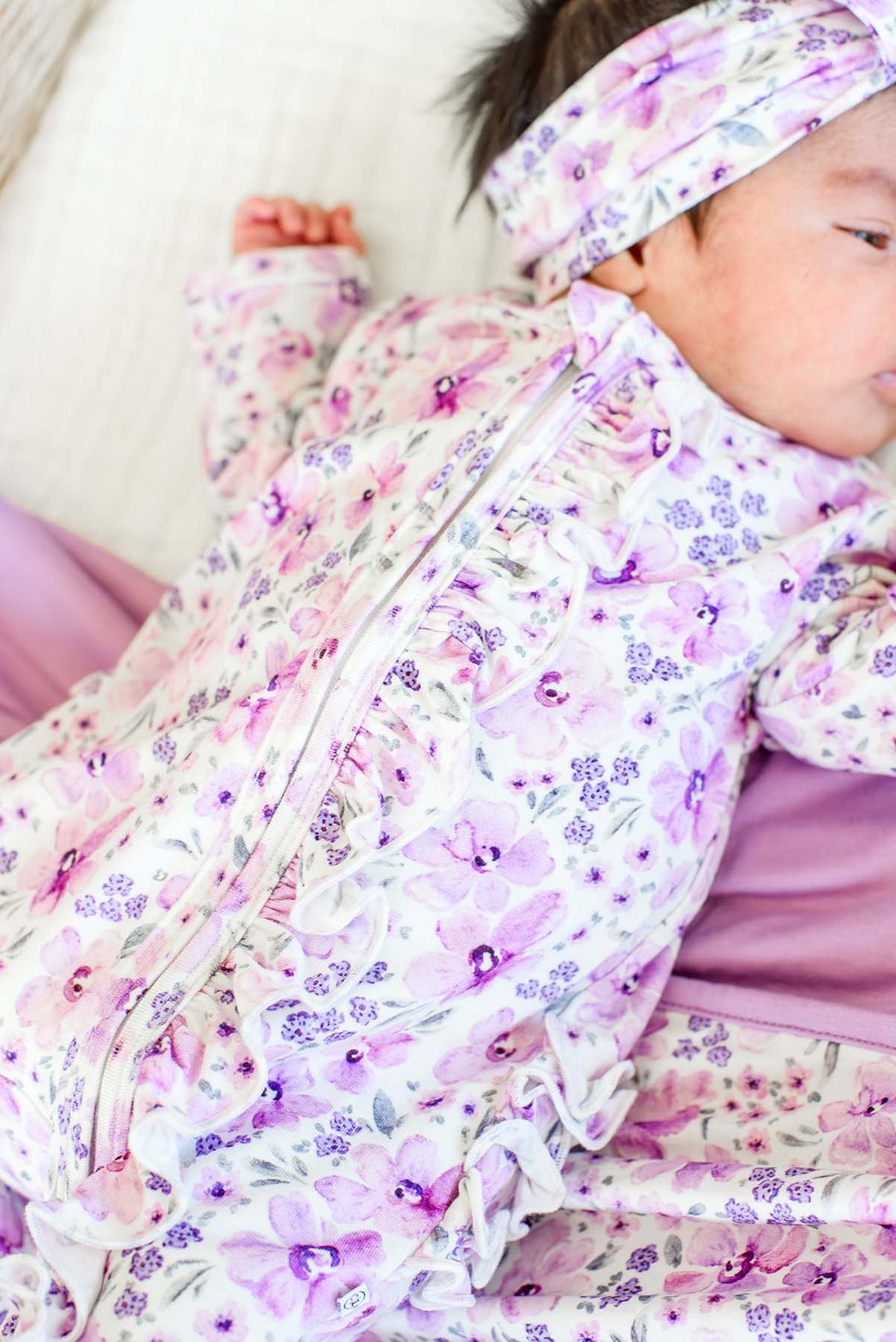 Close-up of the ruffled zipper placket on a newborn wearing a Caden Lane white knit ruffle footie in a Lilac Dreams print featuring lavender florals, paired with a matching headwrap.