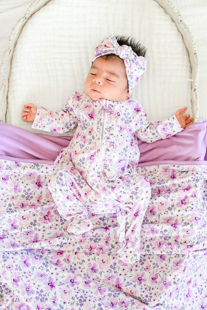 Newborn, arms outstretched in a wicker bassinet, wearing a Caden Lane white knit ruffle footie in a Lilac Dreams print featuring purple flowers, paired with a matching headwrap and blanket.