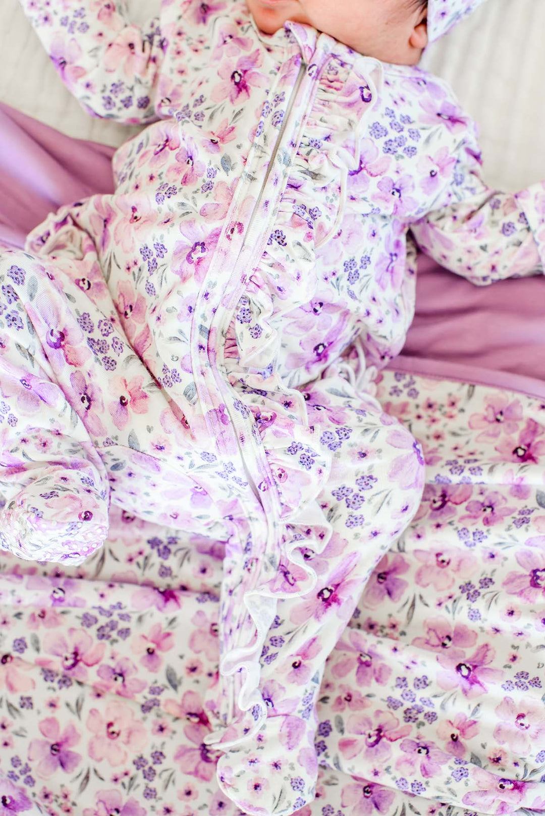Close-up of a newborn, face cropped, wearing a Caden Lane white knit ruffle footie in a Lilac Dreams print featuring purple watercolor flowers, paired with a matching blanket.