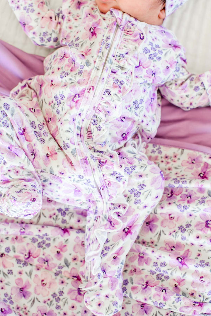 Close-up of a newborn, face cropped, wearing a Caden Lane white knit ruffle footie in a Lilac Dreams print featuring purple watercolor flowers, paired with a matching blanket.