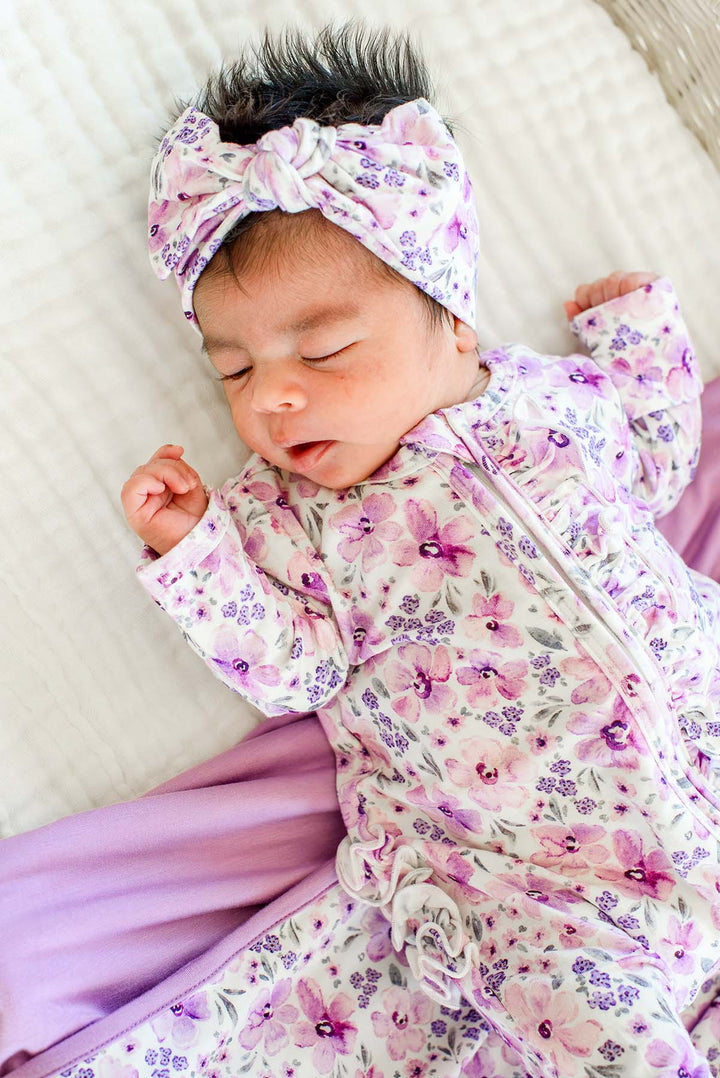 Sleeping newborn with one hand raised, wearing a Caden Lane white knit ruffle footie in a Lilac Dreams print featuring pink watercolor blooms, paired with a matching knotted headwrap.
