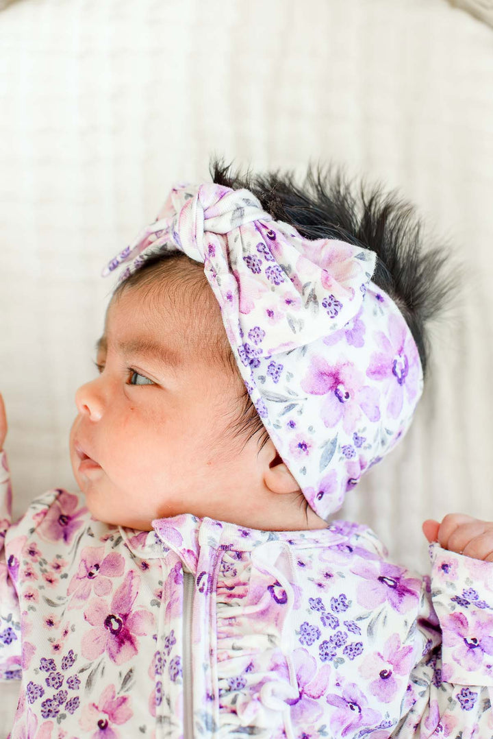 Close-up of a newborn in profile wearing a Caden Lane white knit knotted headwrap in a Lilac Dreams print featuring pink and purple flowers, paired with a matching ruffle footie.