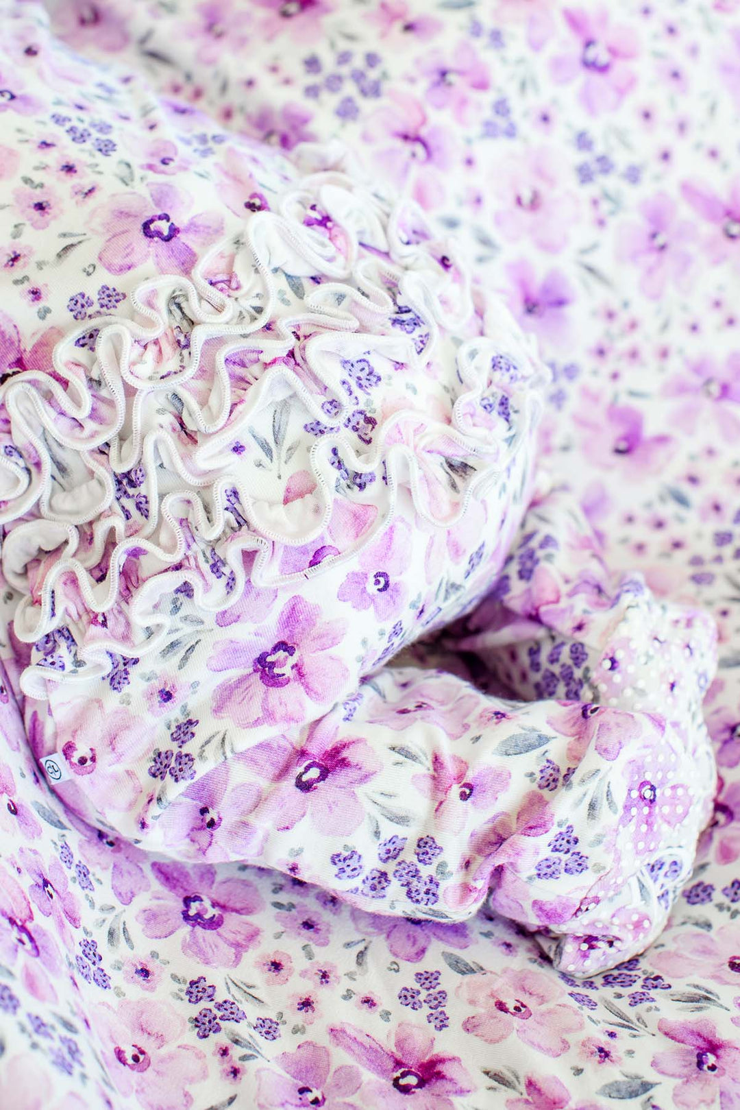 Close-up of the rear ruffles and gripper soles on a Caden Lane white knit ruffle footie in a Lilac Dreams print featuring pink and purple watercolor flowers.