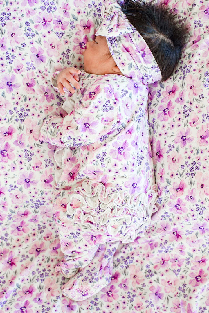Back view of a newborn lying on her side, wearing a Caden Lane white knit ruffle footie in a Lilac Dreams print featuring purple flowers, paired with a matching blanket.