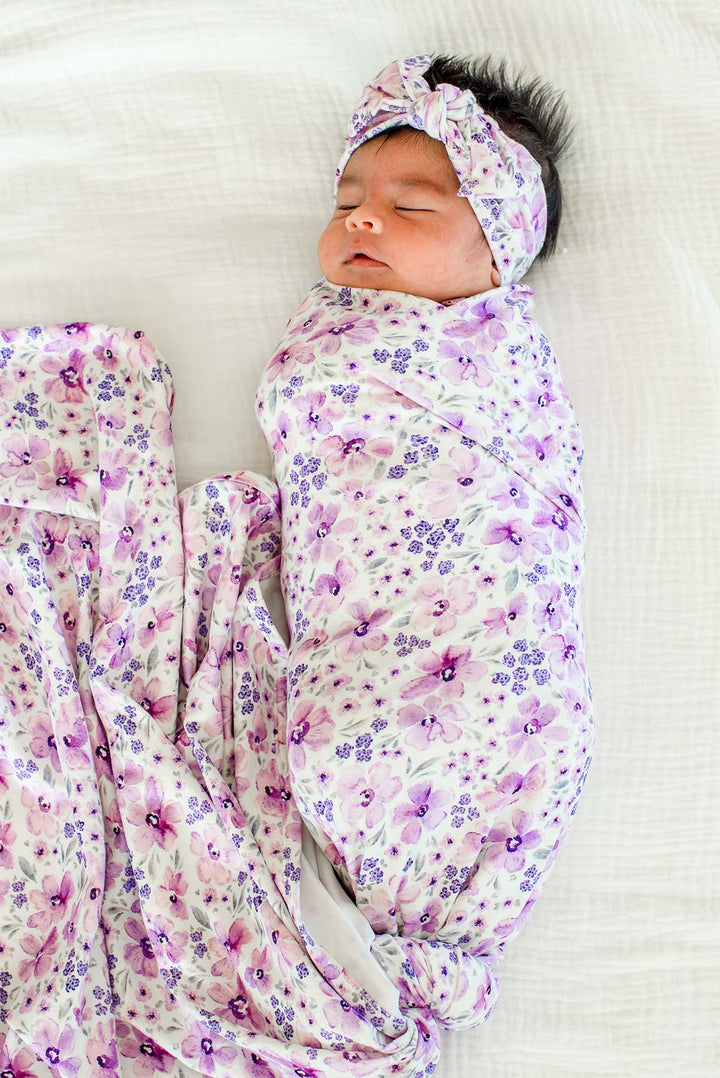 Full-length view of a sleeping newborn wearing a Caden Lane white knit swaddle in a Lilac Dreams print featuring purple flowers and grey leaves, paired with a matching headwrap.