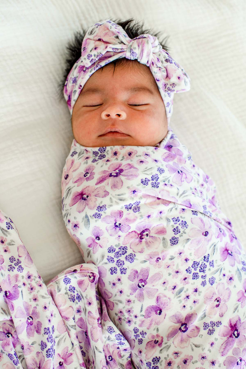 Sleeping newborn facing the camera, wearing a Caden Lane white knit swaddle in a Lilac Dreams print featuring pink watercolor blooms, paired with a matching knotted headwrap.