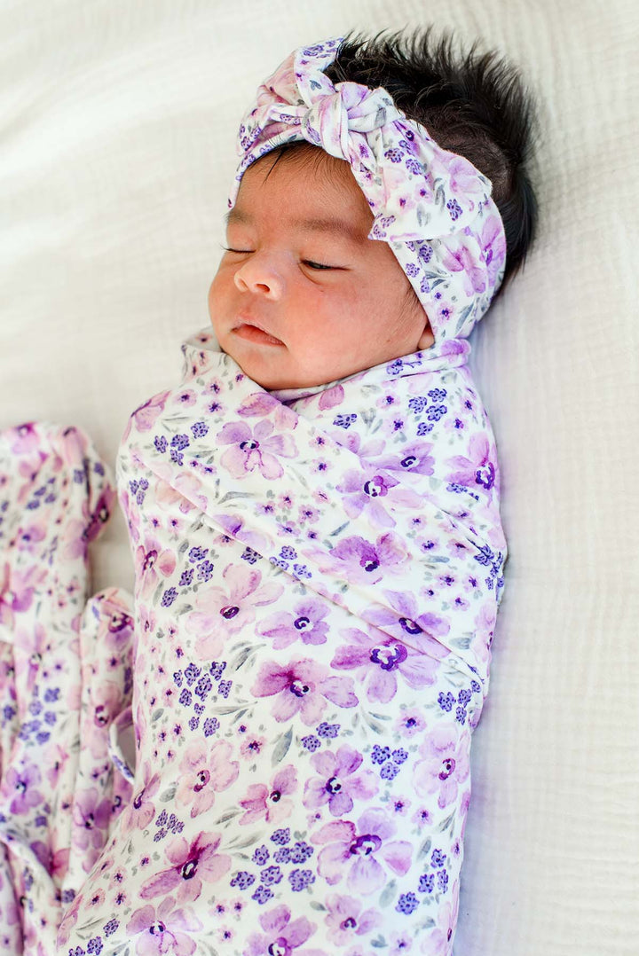Sleeping newborn with her head turned slightly, wearing a Caden Lane white knit swaddle in a Lilac Dreams print featuring tiny violet florals, paired with a matching headwrap.