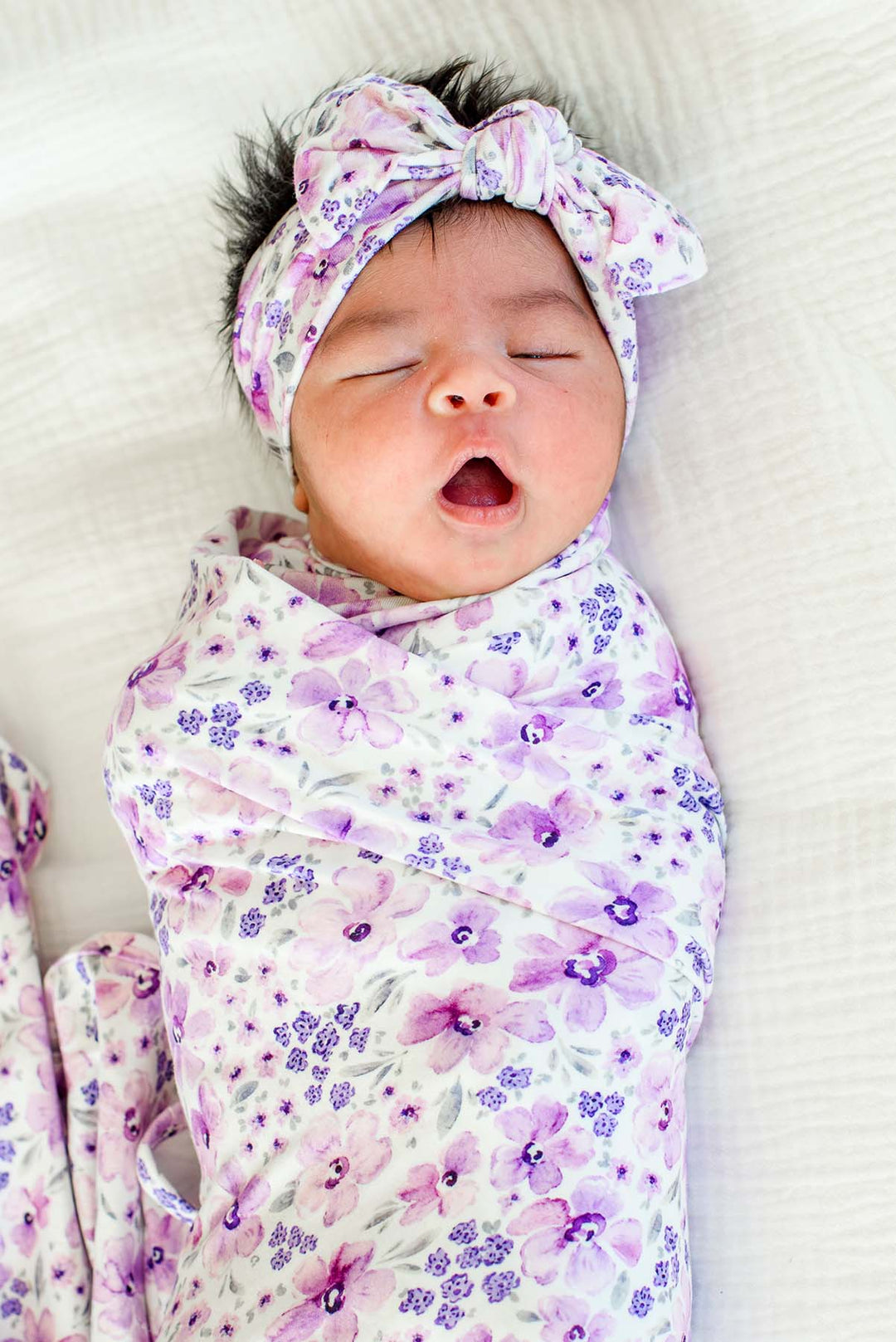 Yawning newborn wearing a Caden Lane white knit swaddle in a Lilac Dreams print featuring lavender watercolor flowers, paired with a matching headwrap tied in a top knot.