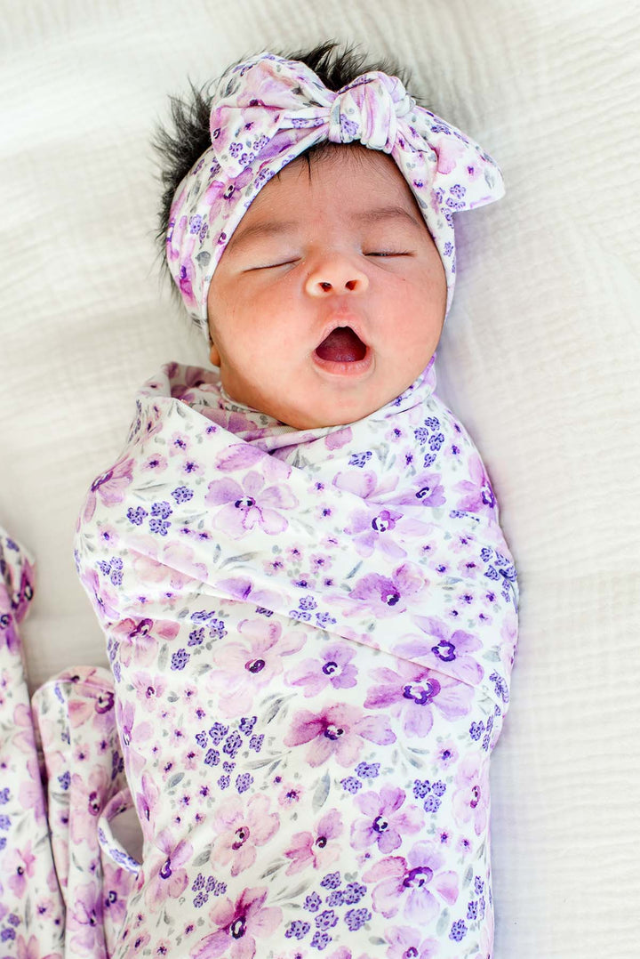 Yawning newborn wearing a Caden Lane white knit swaddle in a Lilac Dreams print featuring lavender watercolor flowers, paired with a matching headwrap tied in a top knot.