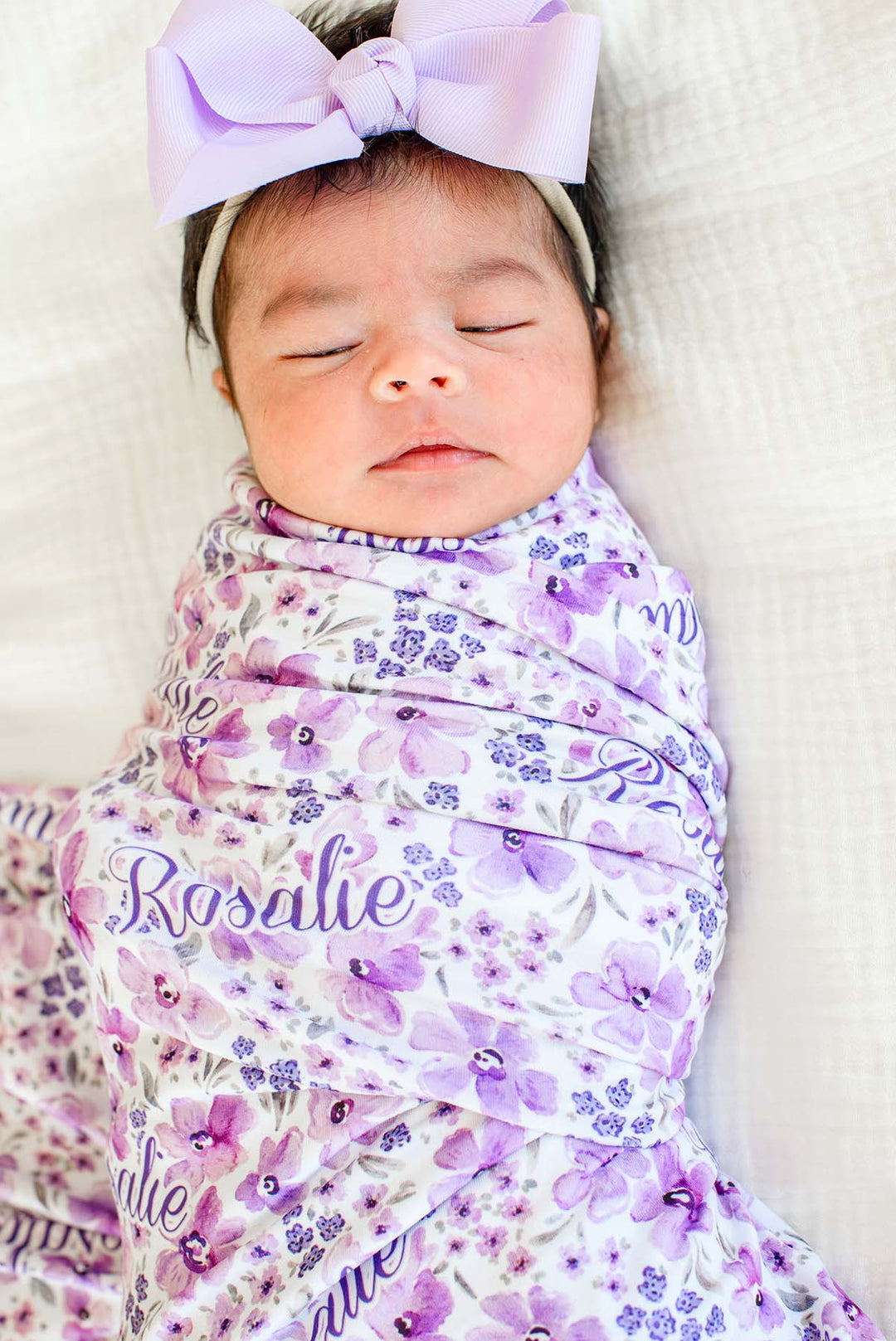 Close-up of a sleeping newborn with a lilac bow headband, wearing a Caden Lane white knit swaddle in a Lilac Dreams print featuring purple watercolor flowers, reading "Rosalie."