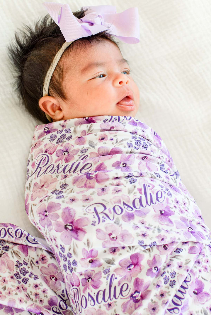 Awake newborn with a lilac bow headband gazing to the side, wearing a Caden Lane white knit swaddle in a Lilac Dreams print featuring violet watercolor flowers, reading "Rosalie."