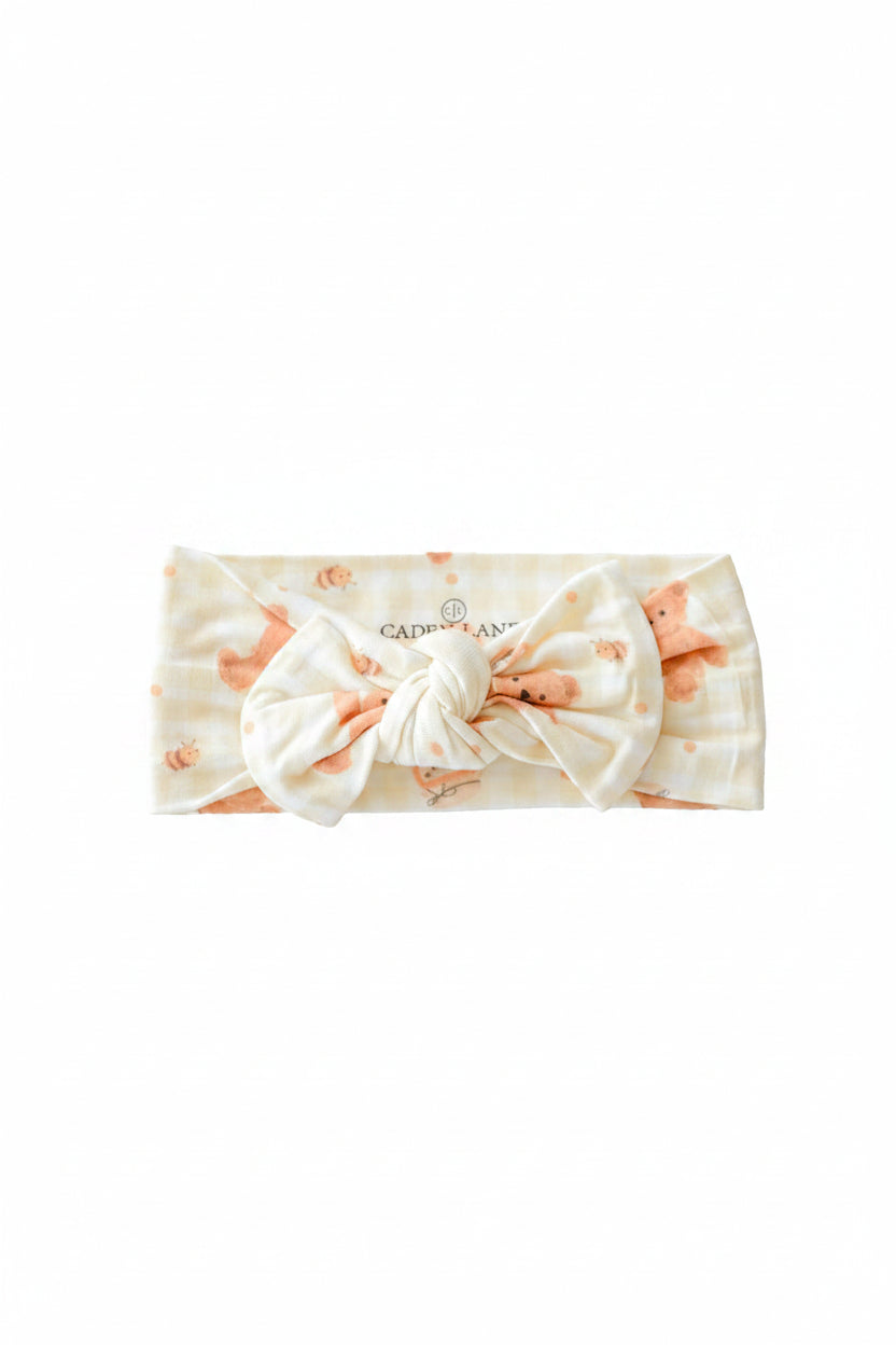 Caden Lane baby headband with a knotted bow in a honey bear and bee print on a gingham background, laid flat on a white surface