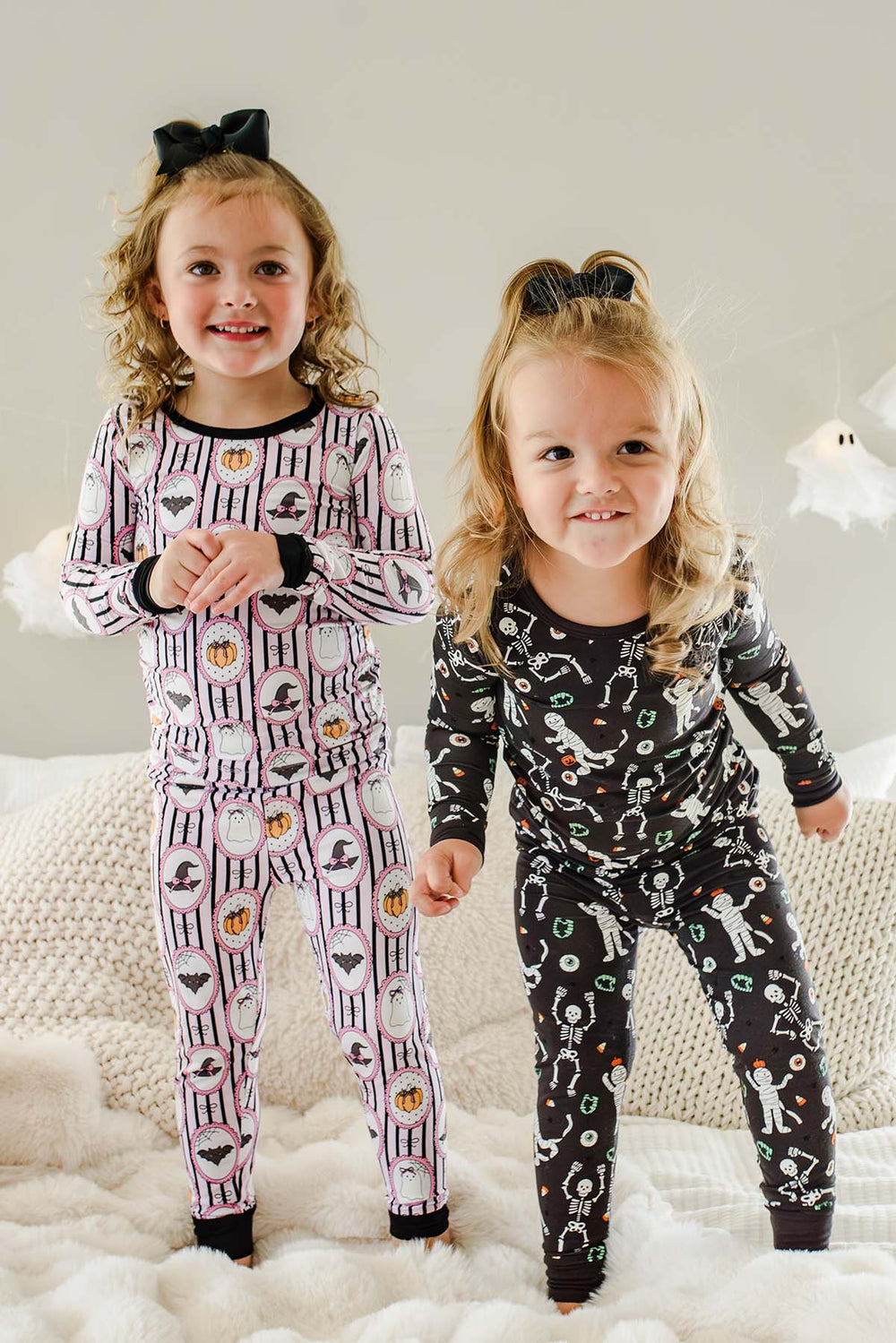 Two young girls wearing matching long-sleeve pajamas with Halloween-themed prints, standing on a white textured blanket in a cozy indoor setting.