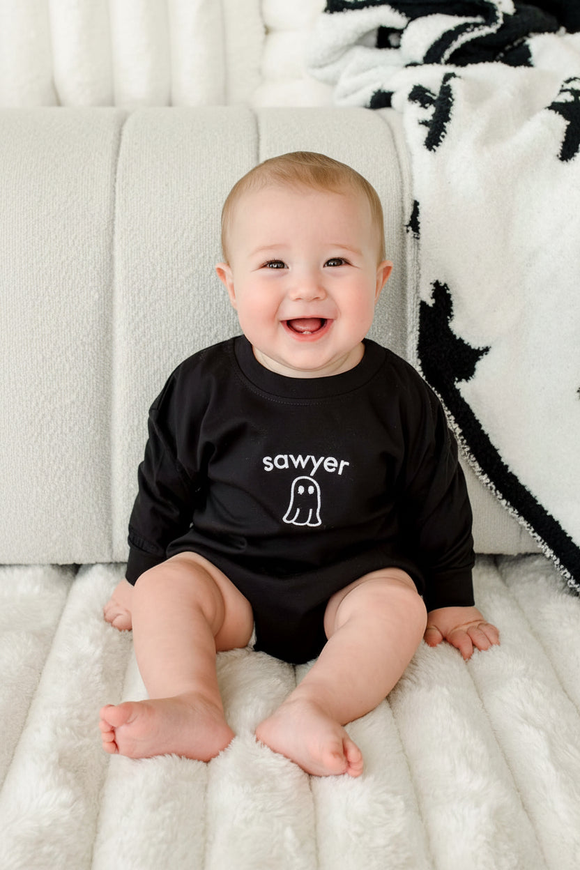 A baby sitting on a white textured blanket in front of a white padded chair, wearing a black long-sleeve shirt with the word 'sawyer' and a small ghost graphic printed in white on the chest.