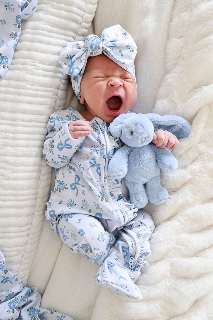 Yawning baby lying down with a stuffed bunny toy, wearing a Tied In Blooms bow headwrap and matching outfit in blue floral and bow print