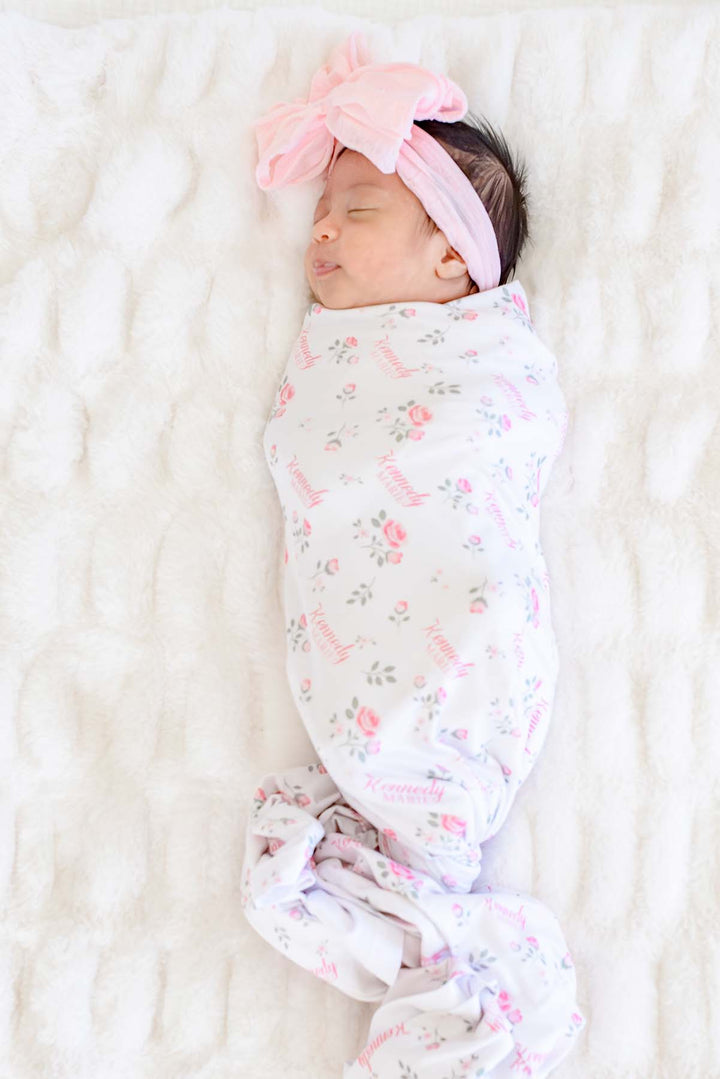 Caden Lane personalized newborn swaddle, sleeping baby swaddled in Rose Garden white floral blanket with custom name and pink bow headband