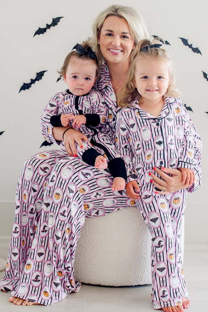 Mother and two daughters wearing matching Caden Lane pink and black striped Halloween pajamas with ghost, bat, and pumpkin cameo print, sitting together against a bat-decorated backdrop.