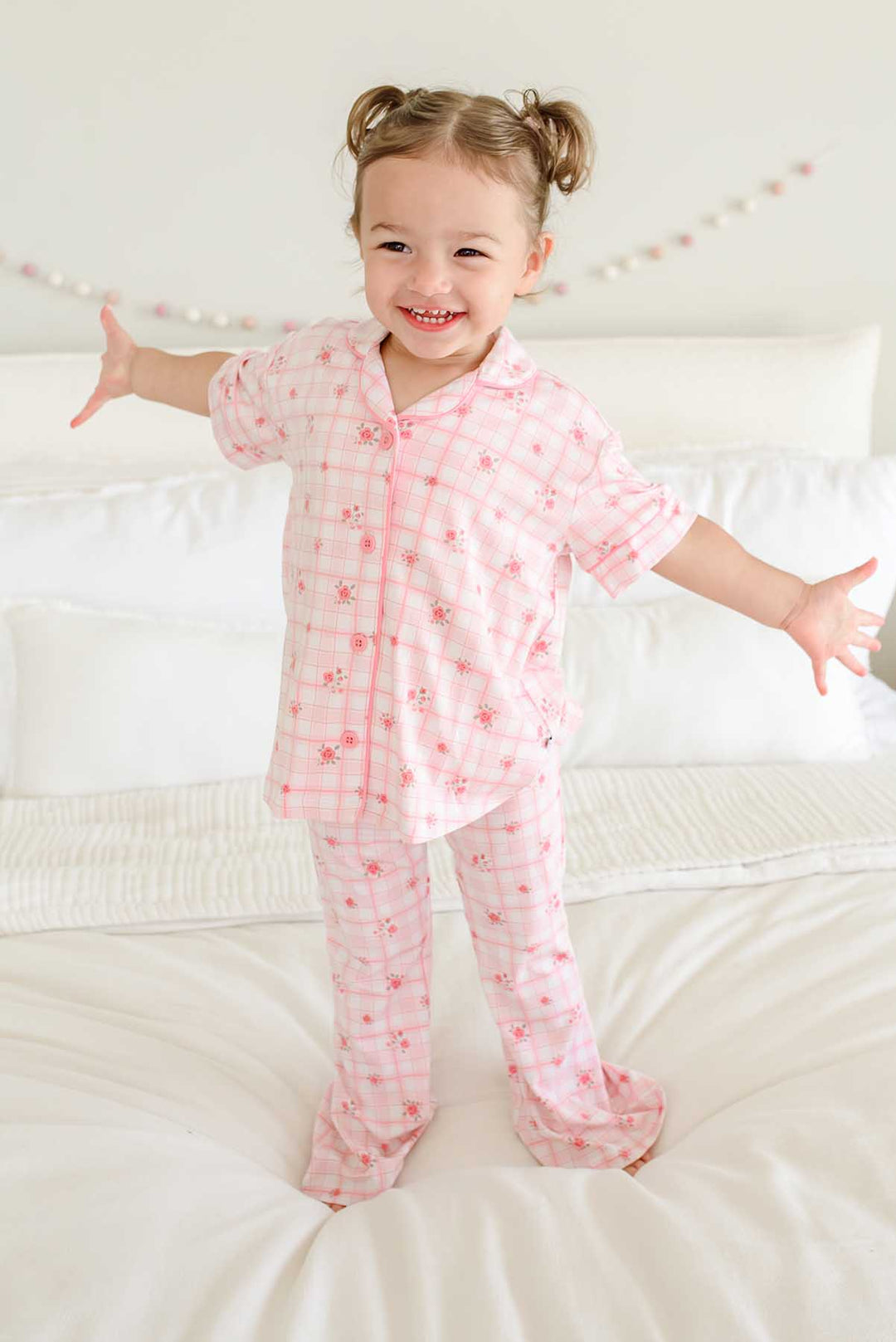 Joyful girl standing on bed with arms open in plaid pajama set