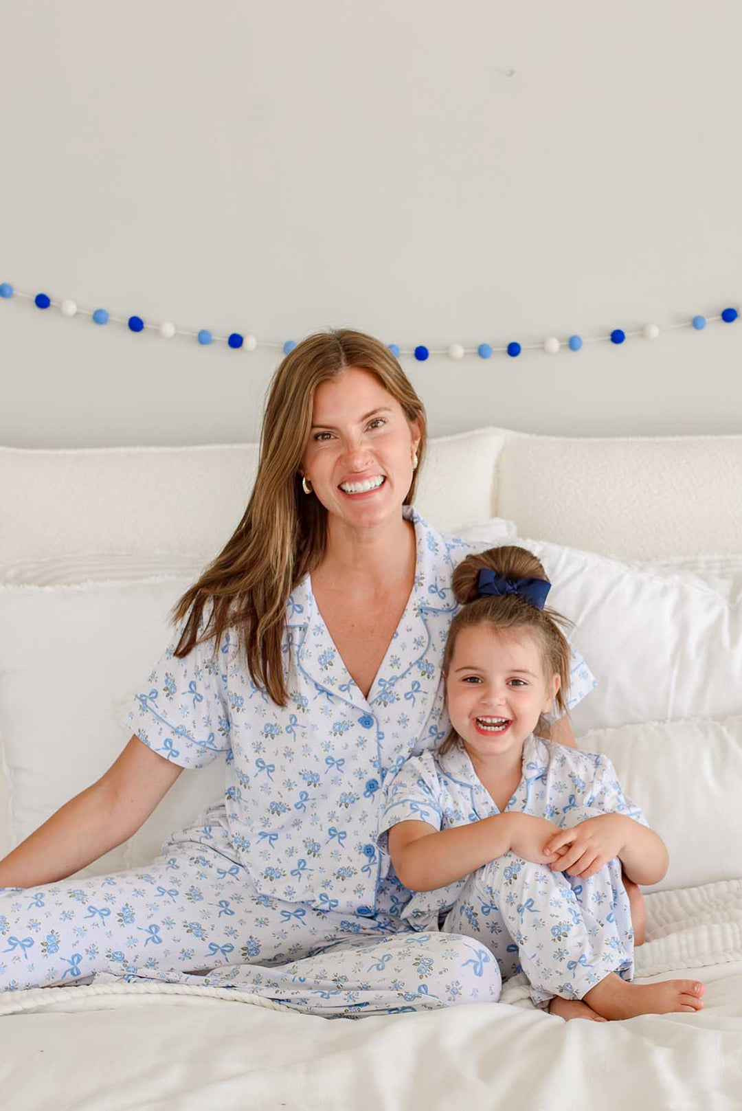 Mother and daughter sitting together on a bed smiling, both wearing matching Tied In Blooms bamboo lounge sets in blue floral and bow print