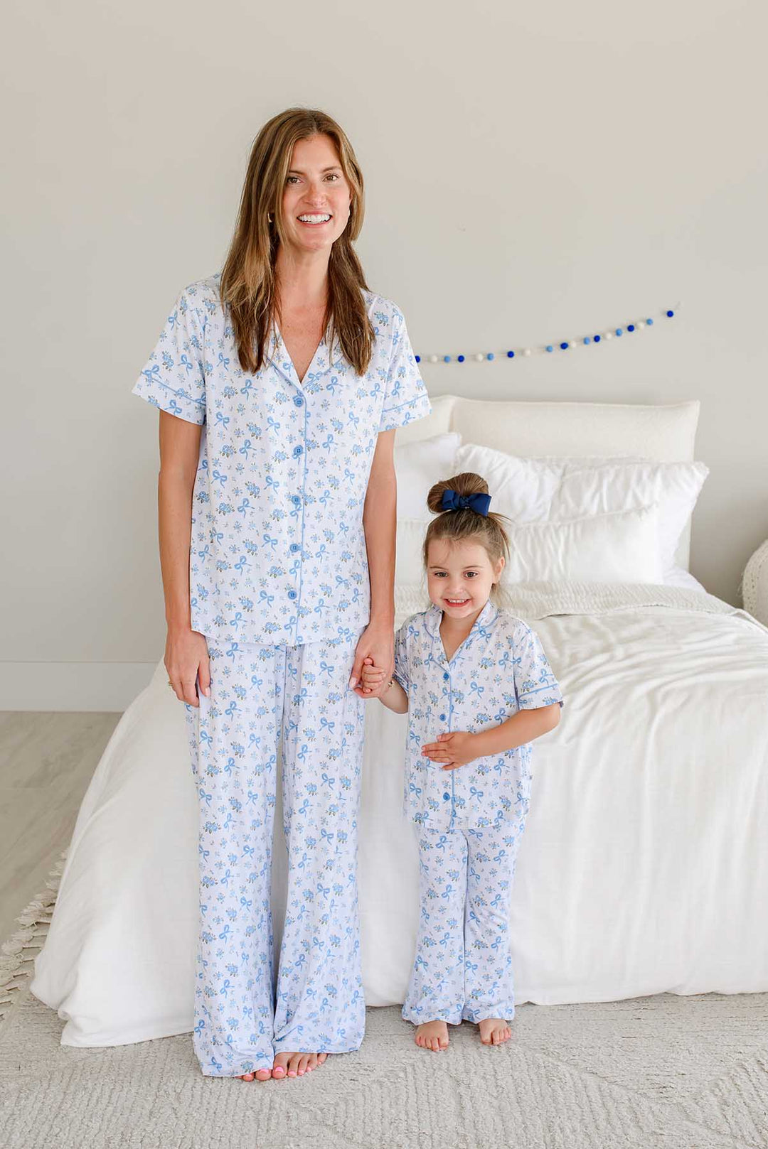 Mother and daughter standing side by side holding hands by a bed, both wearing matching Tied In Blooms bamboo lounge sets in blue floral and bow print
