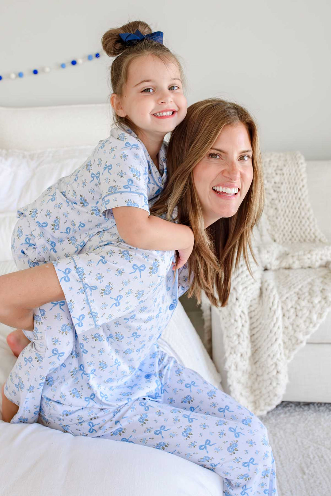 Daughter playfully riding piggyback on her mother, both wearing matching Tied In Blooms bamboo lounge sets in blue floral and bow print