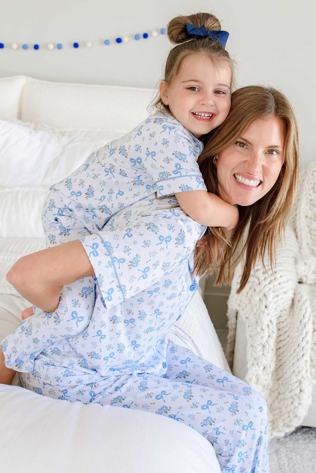 Daughter hugging her smiling mother from behind, both wearing matching Tied In Blooms bamboo lounge sets in blue floral and bow print