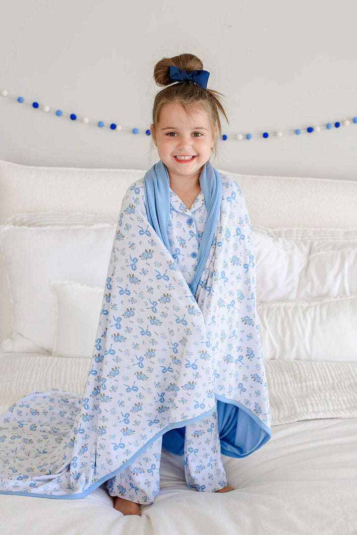 Girl kneeling on a bed and smiling, wrapped in a Tied In Blooms double sided bamboo blanket with blue floral and bow print