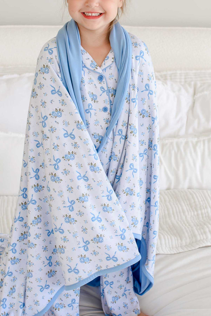 Smiling girl sitting on a bed wrapped in a Tied In Blooms double sided bamboo blanket with blue floral and bow print