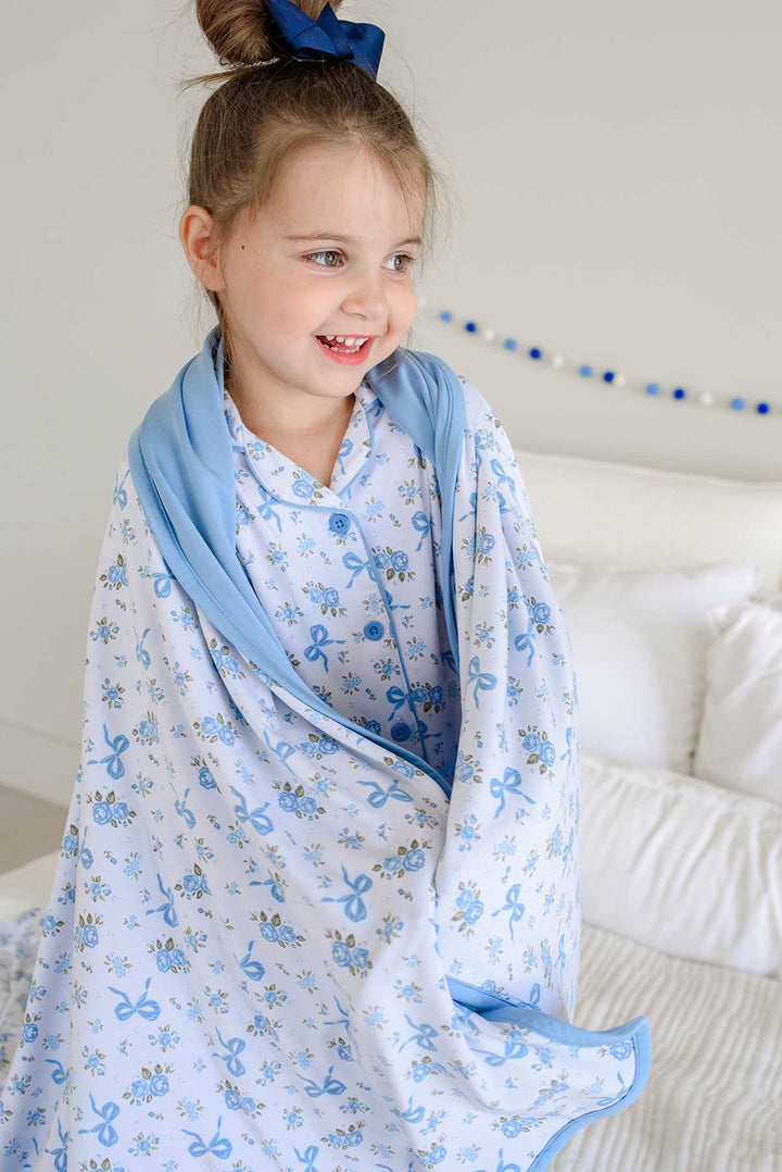 Smiling girl on a bed wrapped in a Tied In Blooms double sided bamboo blanket with blue floral and bow print