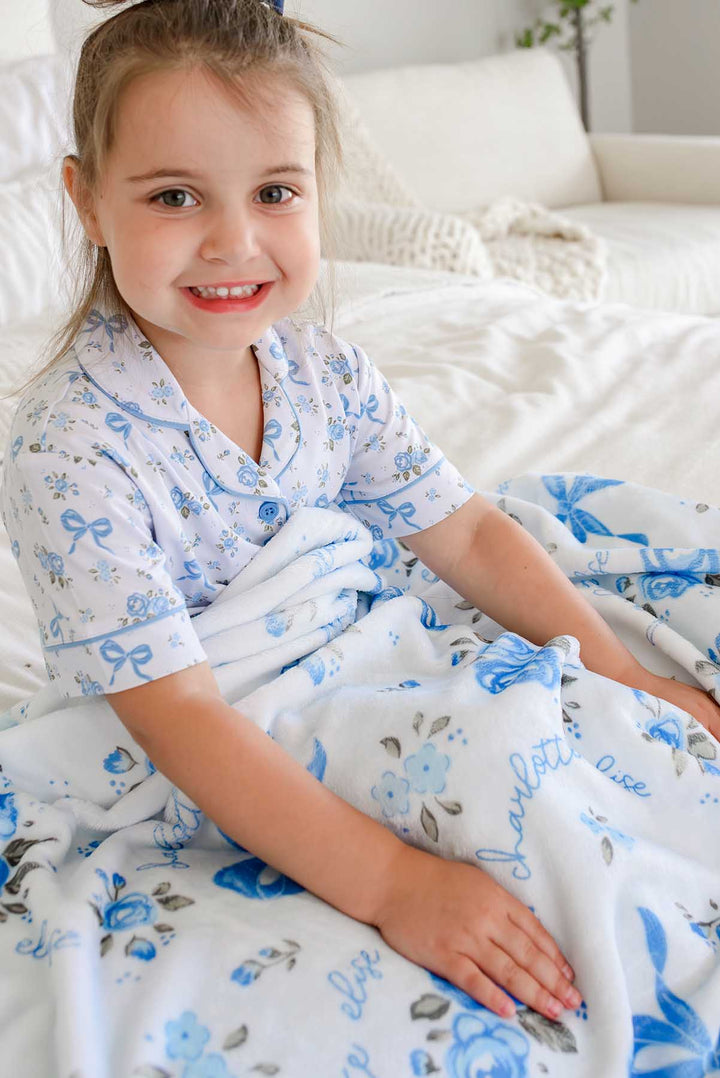 Smiling little girl sitting on a bed in matching pajamas, with a Tied In Blooms personalized blanket in blue floral and bow print draped over her lap