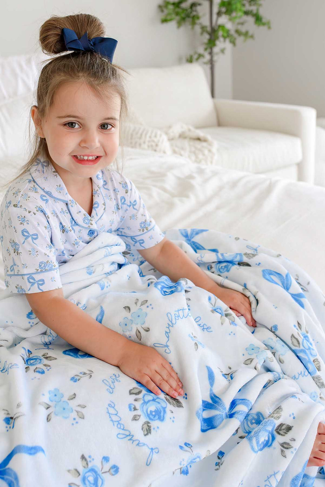 Little girl with a hair bow sitting on a bed in a living room, wrapped in a Tied In Blooms personalized blanket with blue floral and bow print