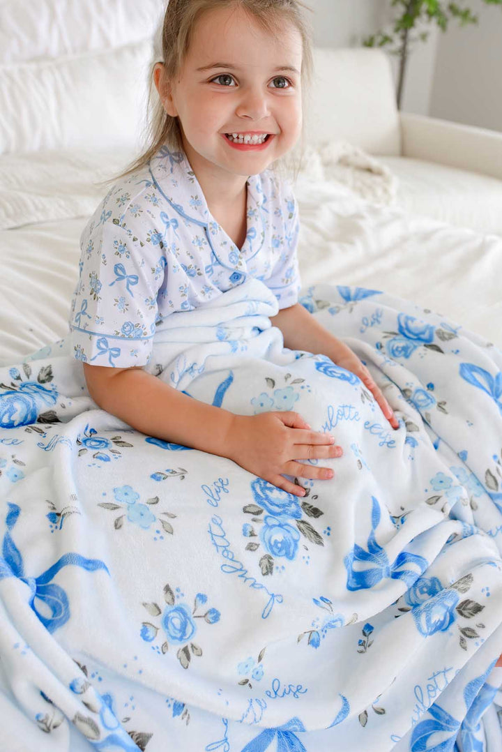 Smiling little girl on a bed with hands on her lap, wrapped in a Tied In Blooms personalized blanket in blue floral and bow print