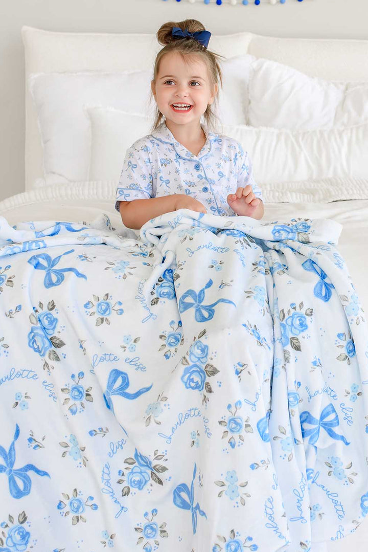 Little girl sitting on a bed smiling, wrapped in a Tied In Blooms personalized blanket with blue floral and bow print and names in cursive script