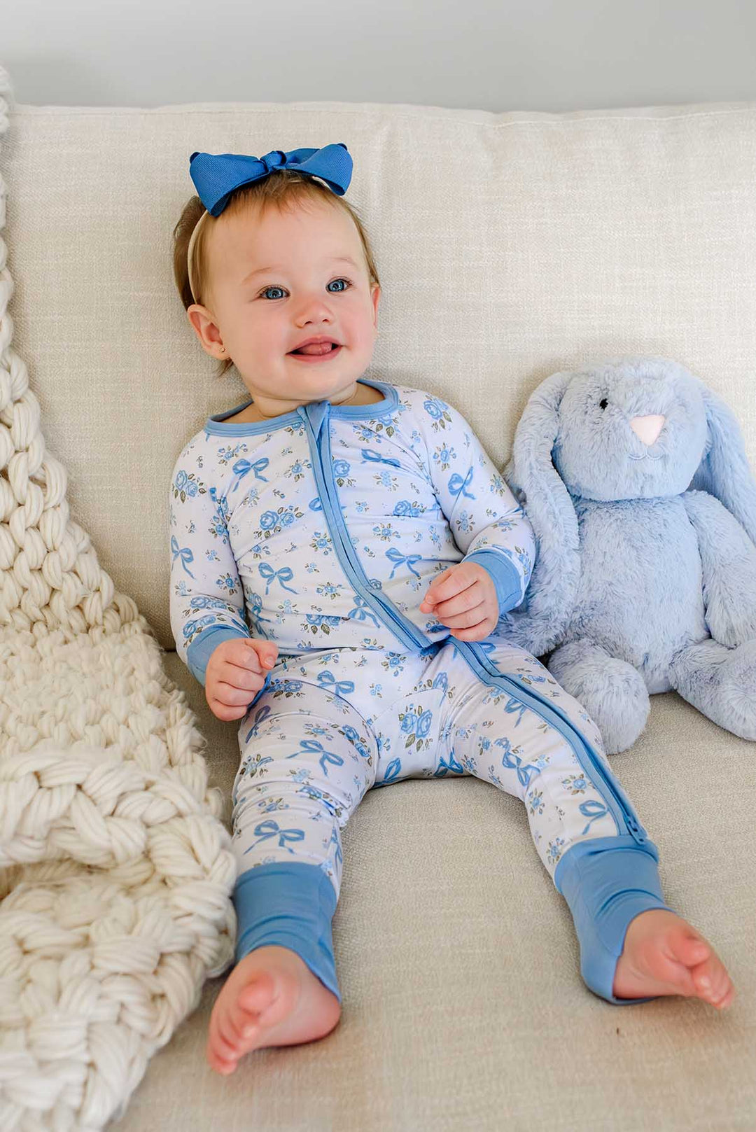 Baby sitting on a couch next to a stuffed bunny toy, wearing a Tied In Blooms convertible bamboo zip romper in blue floral and bow print