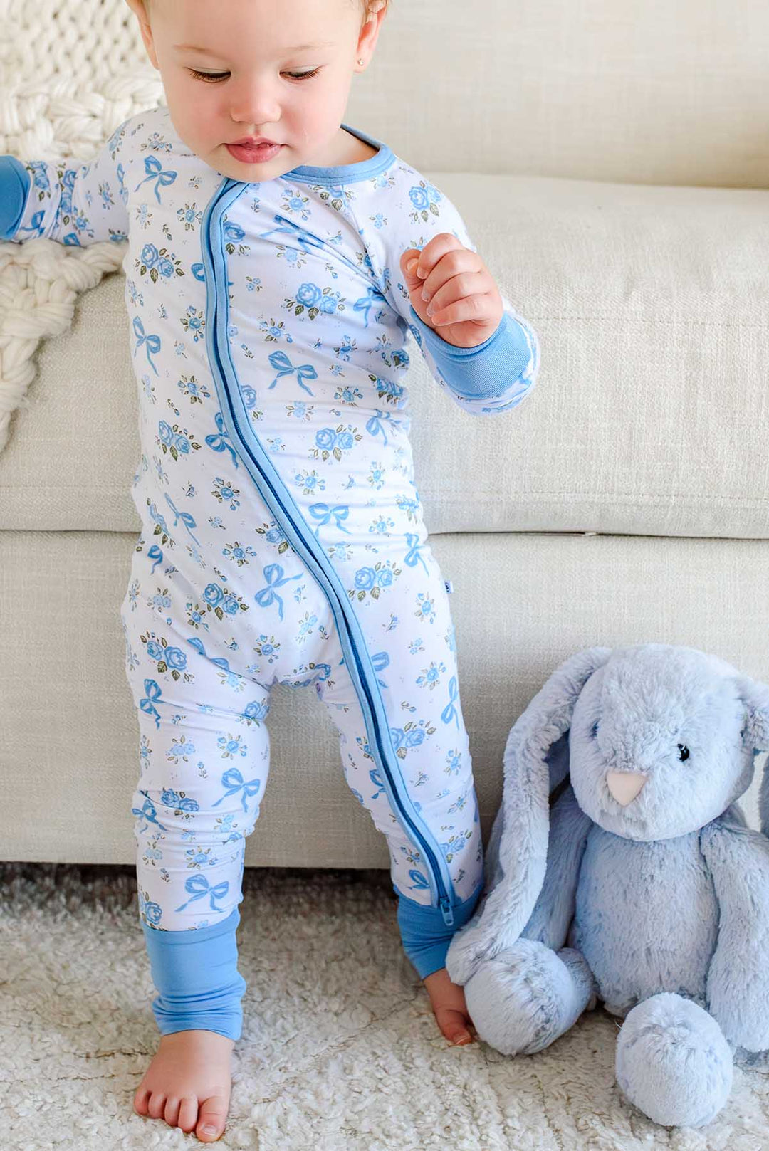 Baby standing next to a stuffed bunny toy, wearing a Tied In Blooms convertible bamboo zip romper in blue floral and bow print