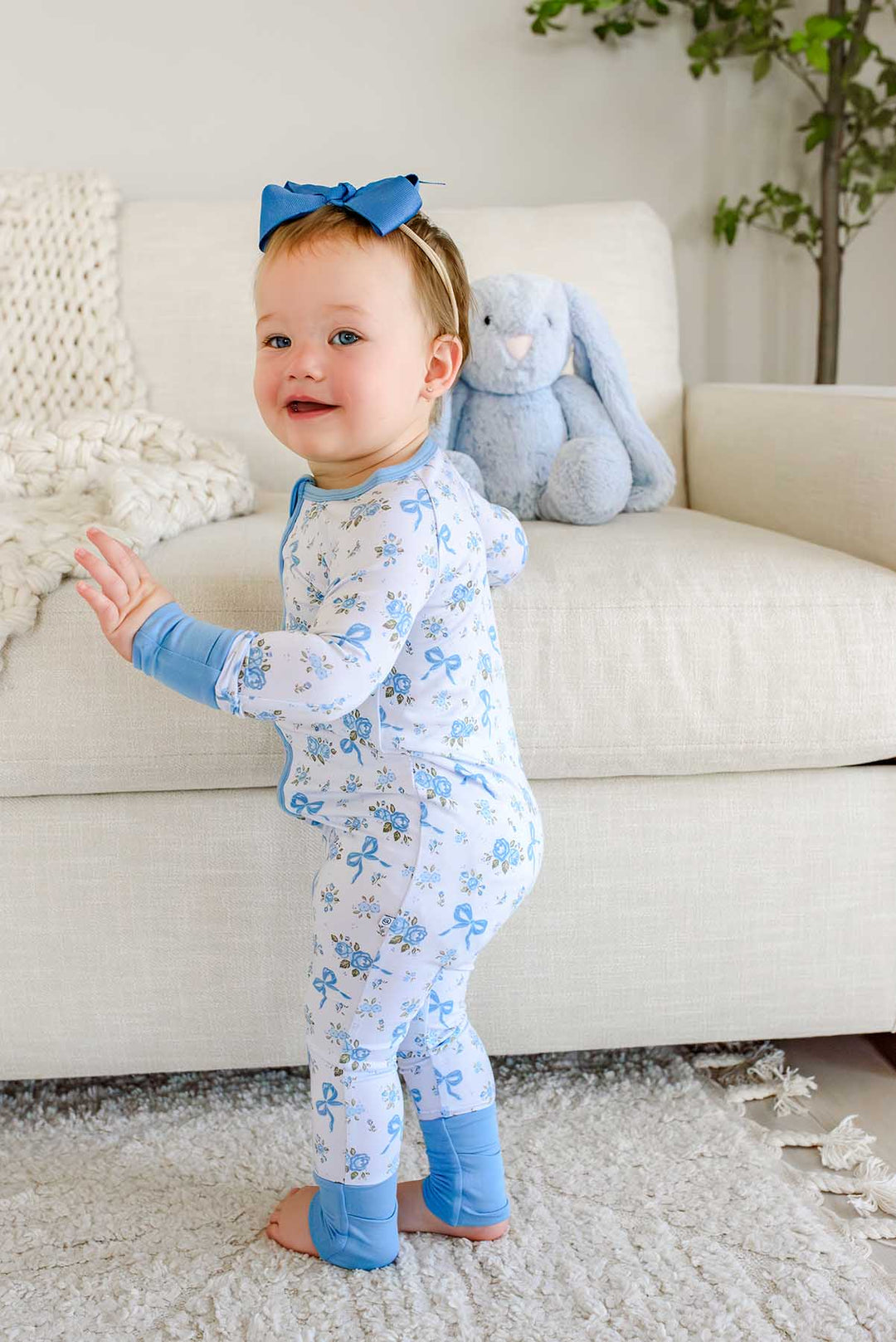Smiling baby standing near a couch and a stuffed bunny toy, wearing a Tied In Blooms convertible bamboo zip romper in blue floral and bow print