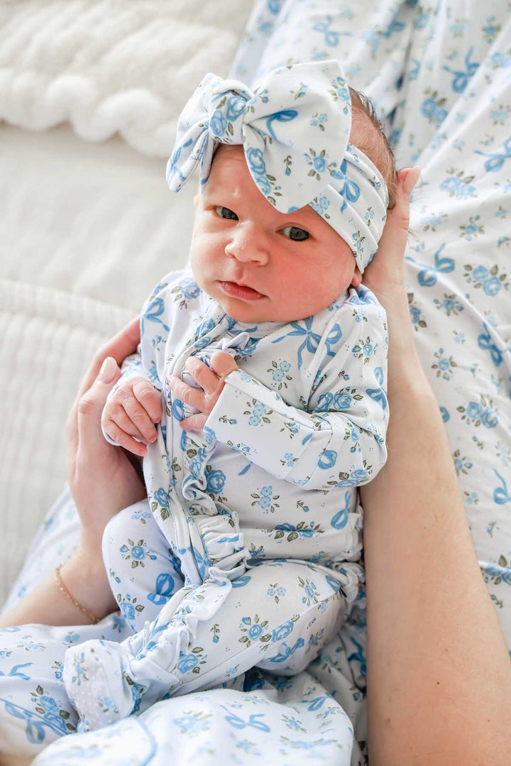 Baby held by an adult, wearing a Tied In Blooms bow headwrap and ruffled outfit in blue floral and bow print