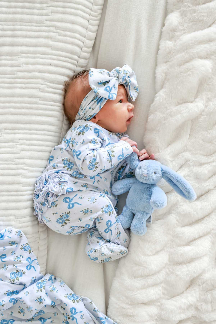 Baby lying with a stuffed bunny toy, wearing a Tied In Blooms bow headwrap and matching outfit in blue floral and bow print