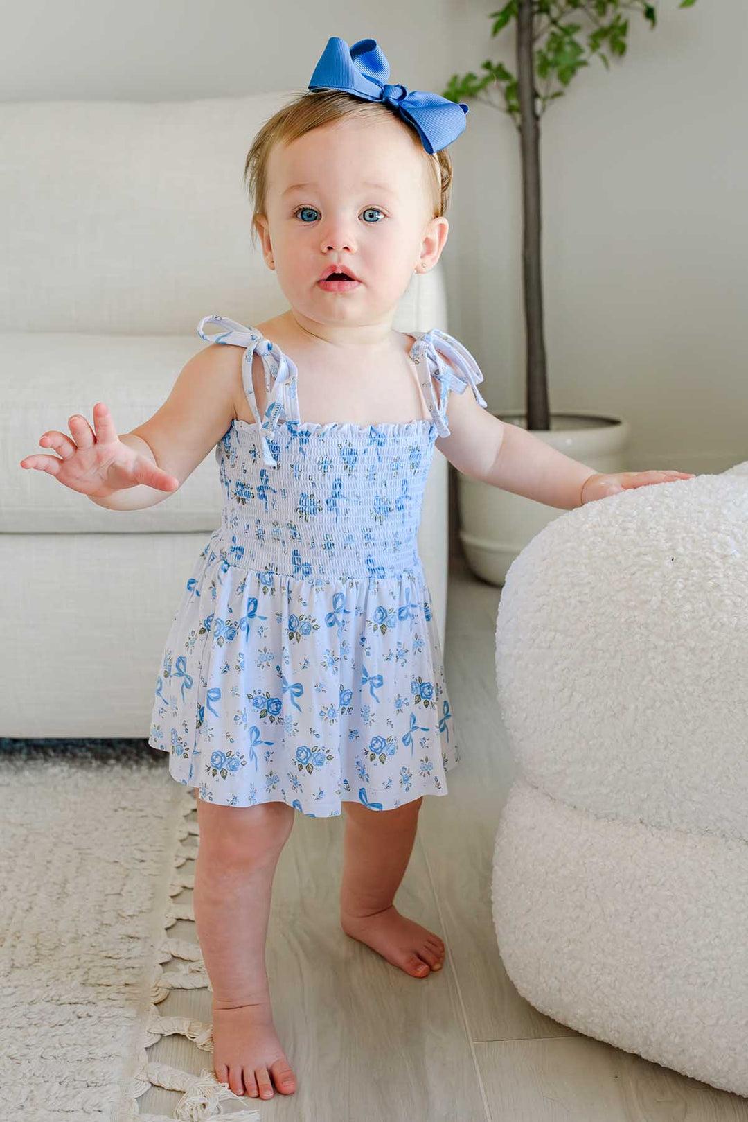 Toddler standing and wearing a Tied In Blooms smocked twirl bodysuit in blue floral and bow print with a matching bow headband