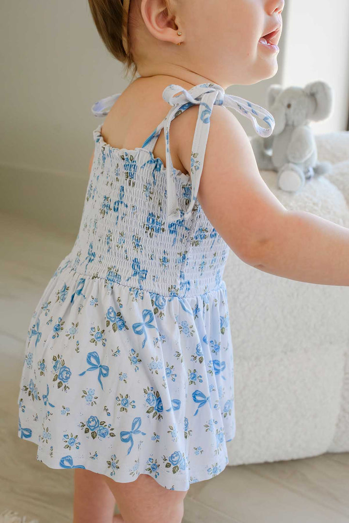 Back view of a toddler near a stuffed elephant toy, wearing a Tied In Blooms smocked twirl bodysuit in blue floral and bow print